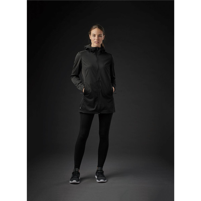 A woman wearing a black Women's Belcarra Softshell jacket, paired with black leggings and sneakers. The jacket features a hood and pockets.