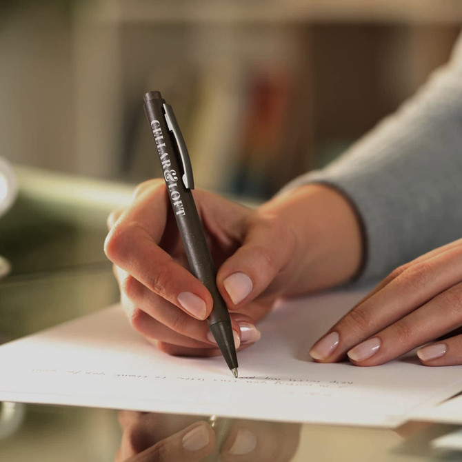 A black pen held by a person, writing on a sheet of paper, with a blurred background. The pen features a logo.
