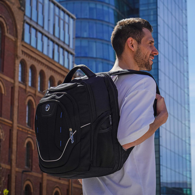 A black backpack with multiple compartments and a logo, worn on a man's shoulder in an urban setting.