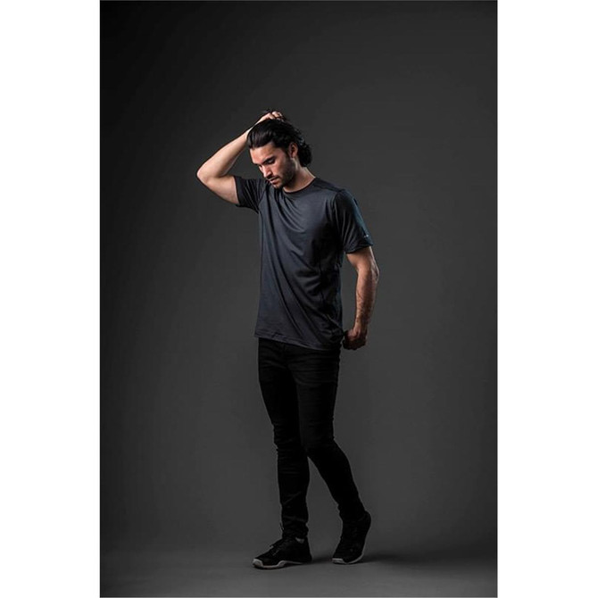 A model wearing a short-sleeve, dark grey T-shirt paired with black pants against a dark background. The shirt features a logo.