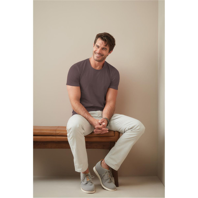 A model sits on a bench, wearing a mauve crew neck t-shirt and light-coloured trousers with grey sneakers.