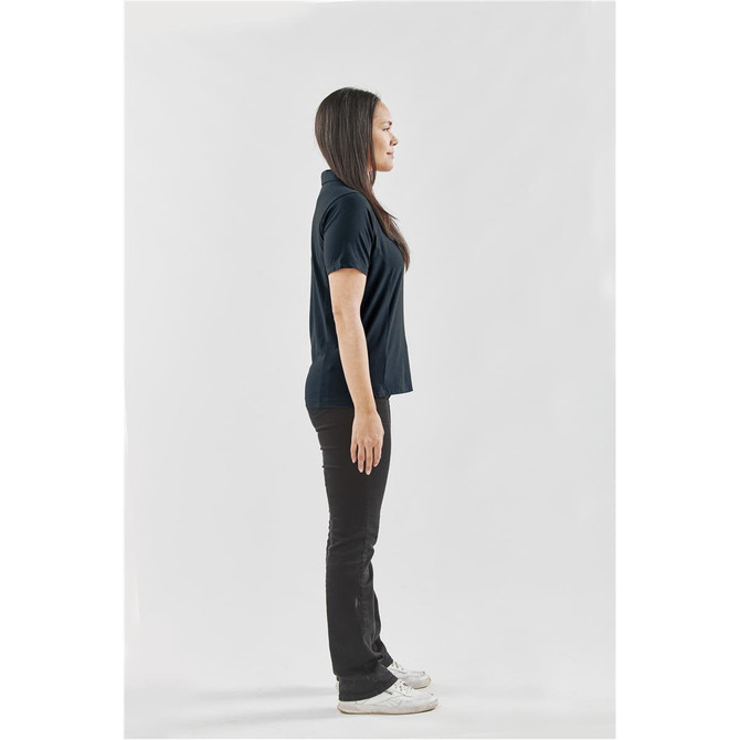 A women's navy polo shirt is displayed on a model standing sideways, paired with black trousers and sneakers.