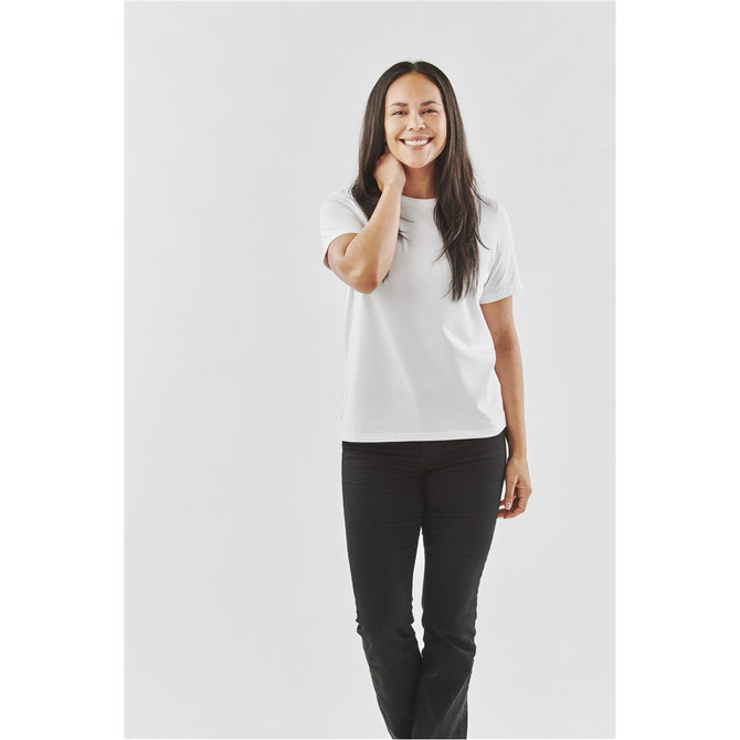 A woman stands smiling in a white Women's Oasis Tee, paired with black pants, against a plain background.