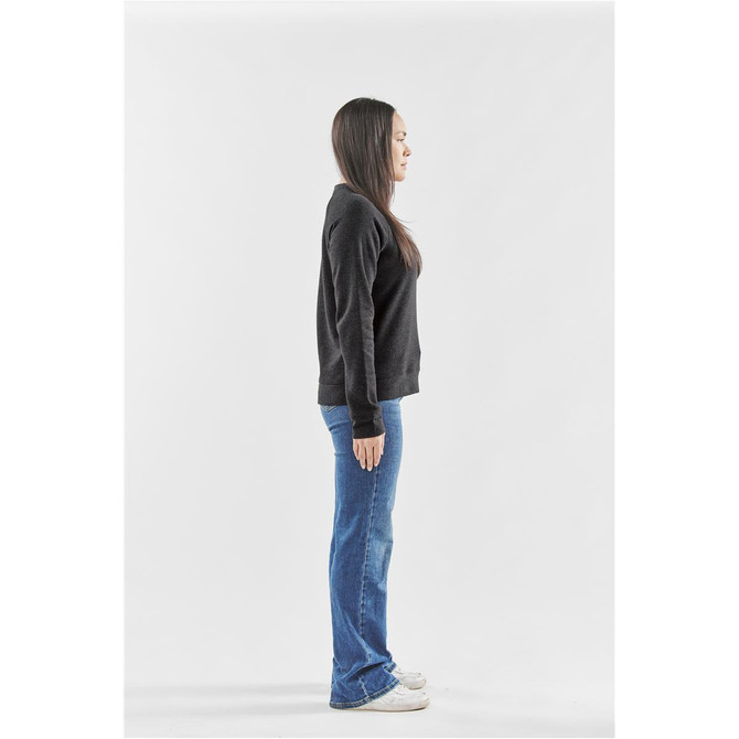 A woman stands sideways, wearing a dark grey crew neck fleece jumper and blue flared jeans, with white sneakers.