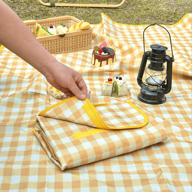 A folded yellow and white checkered picnic blanket lies on grass, accompanied by a lantern and picnic items.