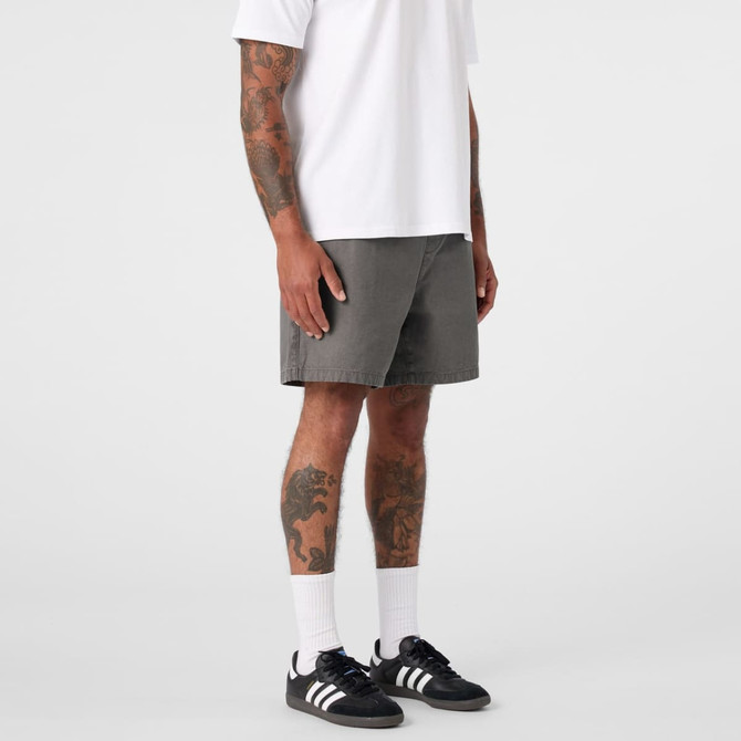 A man stands in grey dress shorts, paired with a white t-shirt and black sneakers, showing his tattoos.