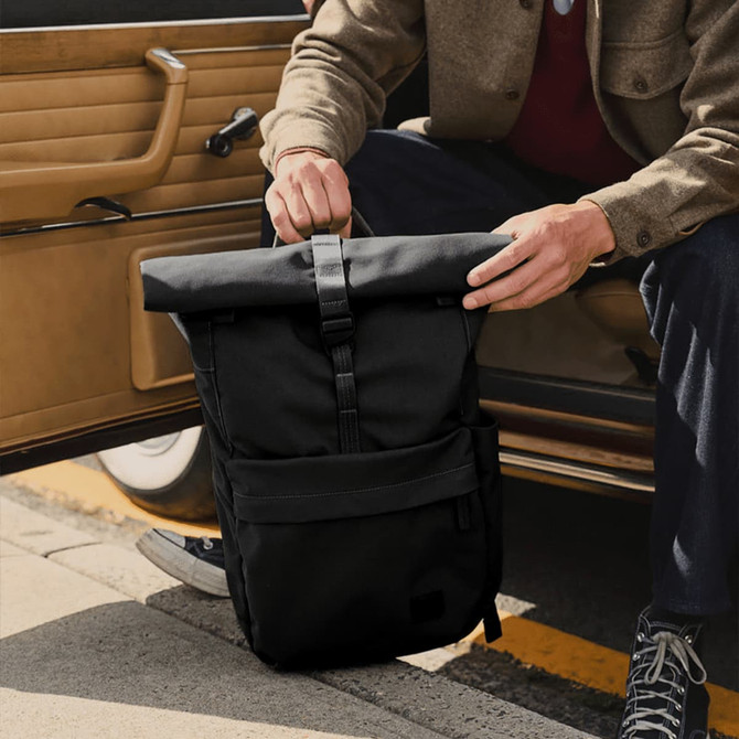 A black rolltop backpack with a front pocket, held by a person sitting in a car. The bag has a logo on it.