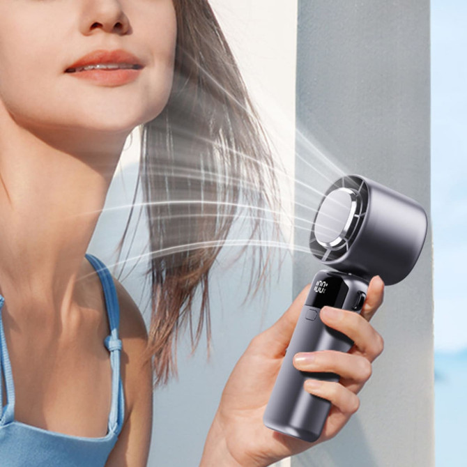 A grey portable cooling fan is held near a person's neck, with mist blowing from the fan. The background is light.