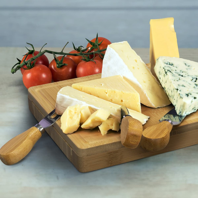 A square wooden cheese board with various cheeses, accompanied by cherry tomatoes and knives with wooden handles.