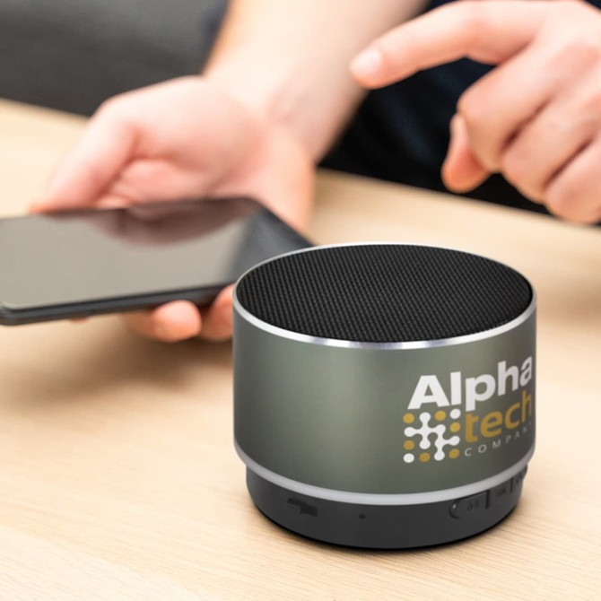 A Bluetooth speaker in grey with a mesh top, featuring a logo, positioned on a table next to a smartphone.