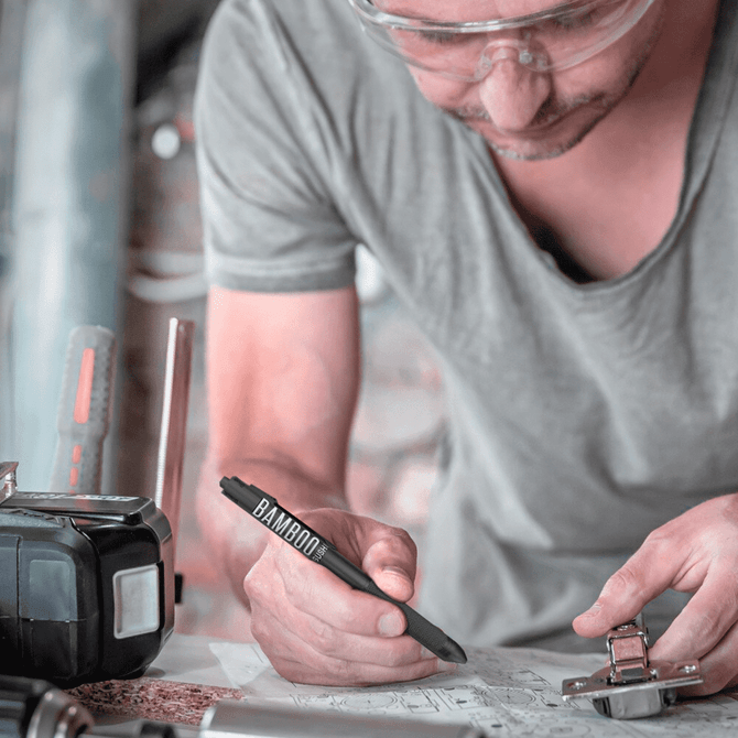 A black metal pen being used by a person while writing on a piece of paper, with tools nearby.