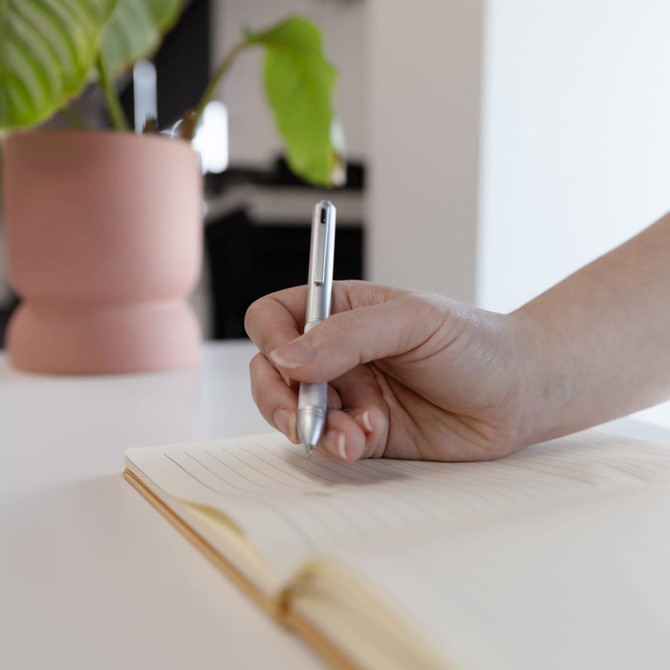 A hand holds a silver metal pen over a lined notepad, with a potted plant in the background.