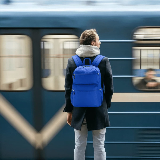 A man stands at a train station wearing a blue High Sierra Everclass Backpack. The background features a blurred train.