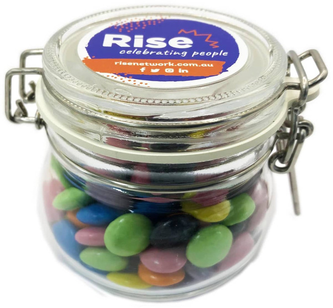 A glass canister filled with colourful choc beans, featuring a metal clasp and a branded lid.