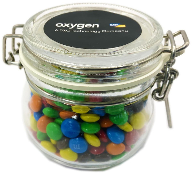 A glass canister filled with colourful mini M&Ms, featuring a metal clasp and a logo on the lid.