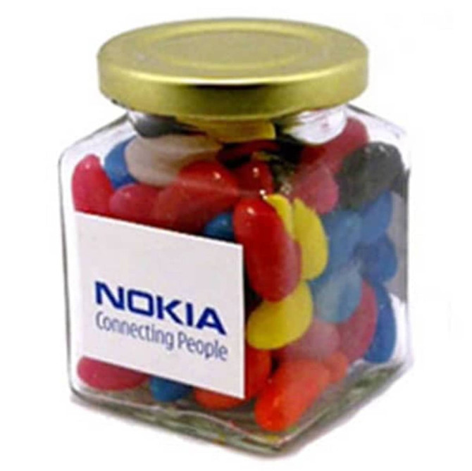A glass square jar filled with colourful mini jelly beans, featuring a gold lid and a logo on the jar.