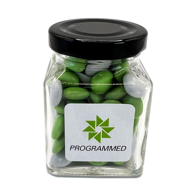 A glass jar containing green and grey choc beans, capped with a black lid and featuring a logo.