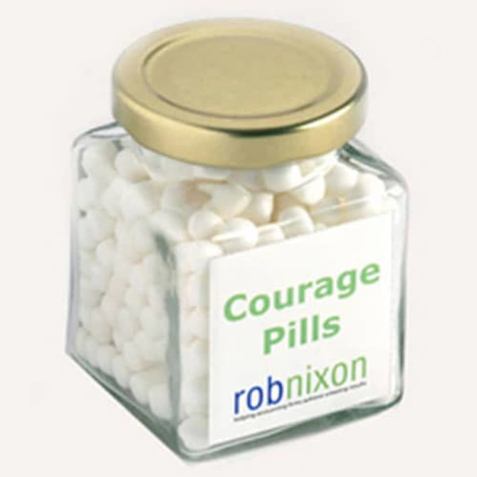 A square glass jar with a gold lid, filled with white mints. The jar features a label with a logo.