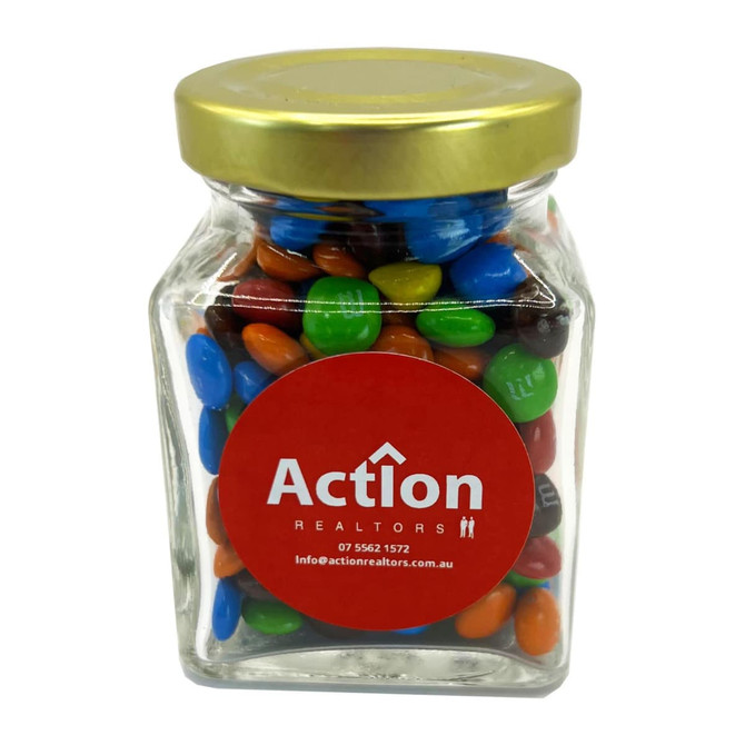 A small glass jar filled with colourful M&Ms in blue, green, red, and orange, topped with a gold lid.