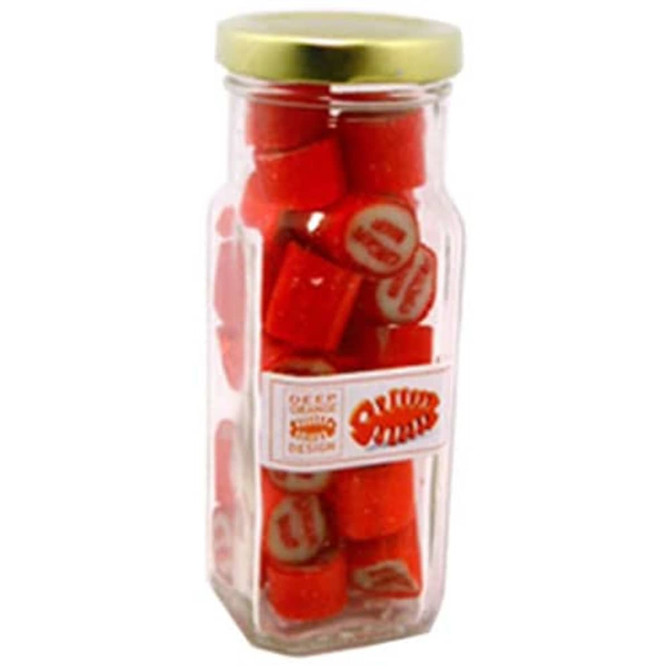 A tall glass jar containing red rock candy pieces with a gold lid and a branding label on the side.