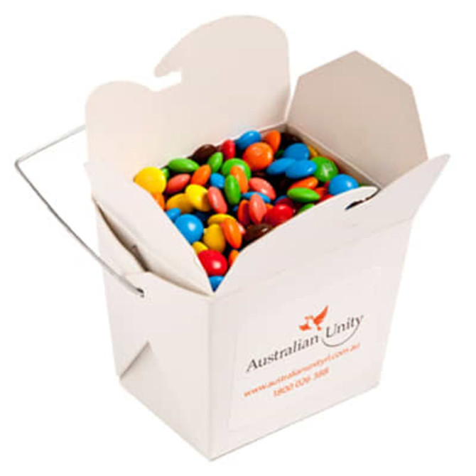 A white cardboard noodle box filled with colourful M&Ms, featuring a metal wire handle.