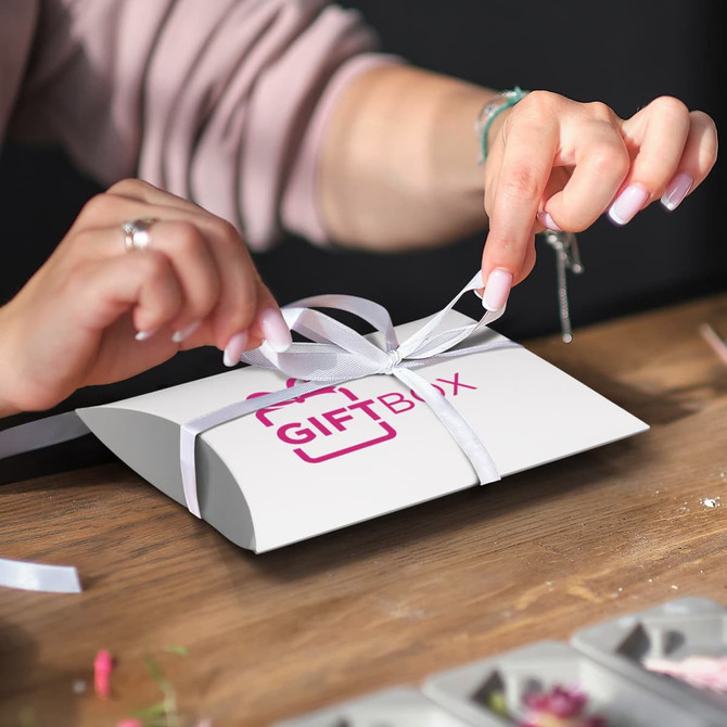 A medium white pillow box with a logo, being tied with a ribbon by a person at a wooden table.