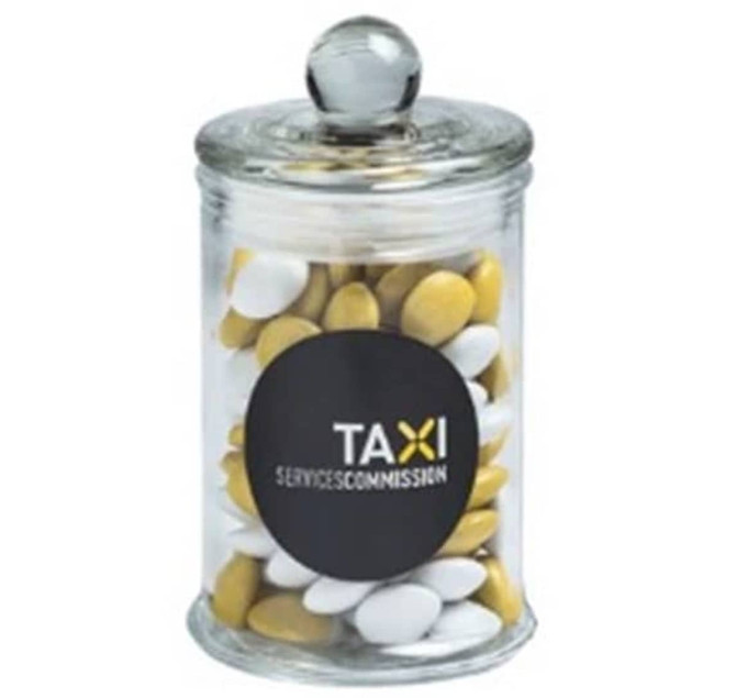 A clear glass apothecary jar filled with yellow and white chocolate beans, featuring a round black logo.