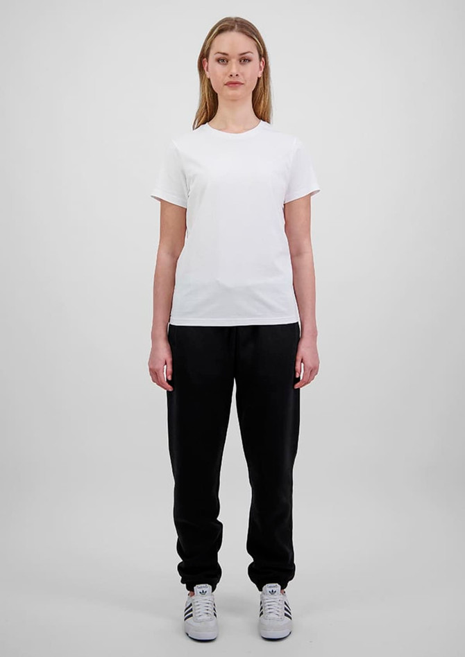 A woman in a white t-shirt and black track pants stands against a plain background. The pants have a branding logo.