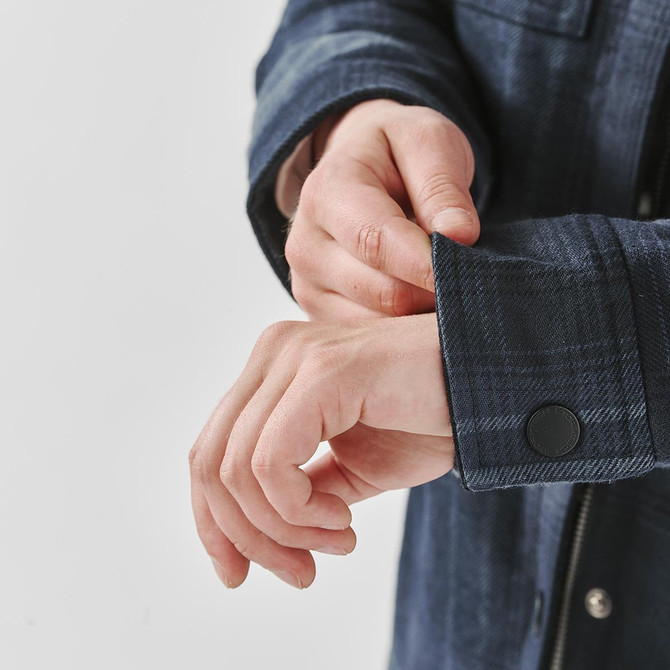 A plaid jacket in a dark blue hue, featuring a Sherpa lining and buttoned cuffs, is being showcased with hands adjusting the sleeve.