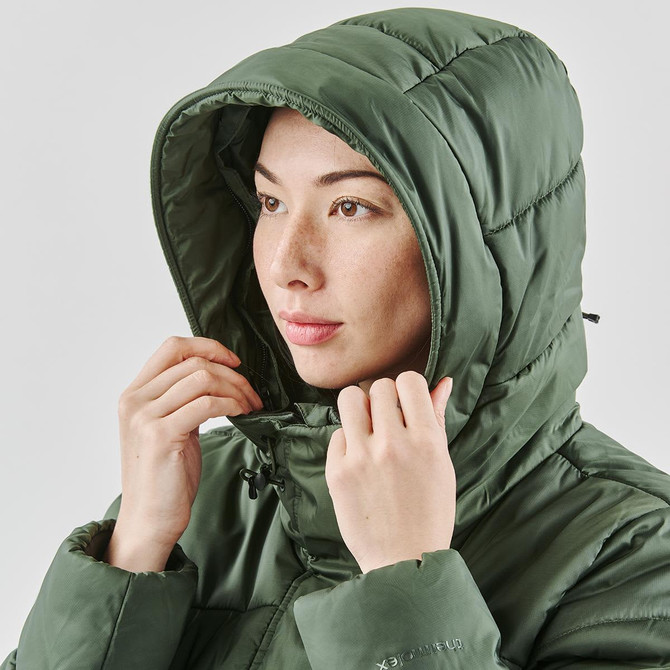 A woman wearing a green puffer jacket with a hood, adjusting it around her face. The jacket has a subtle logo.