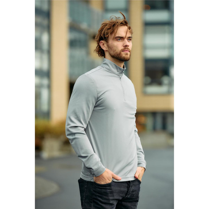 A man wearing a light grey 1/4 zip pullover, standing outdoors with a modern building in the background.