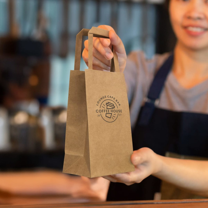 A small brown paper bag with flat handles and a logo, held by a person in an apron.