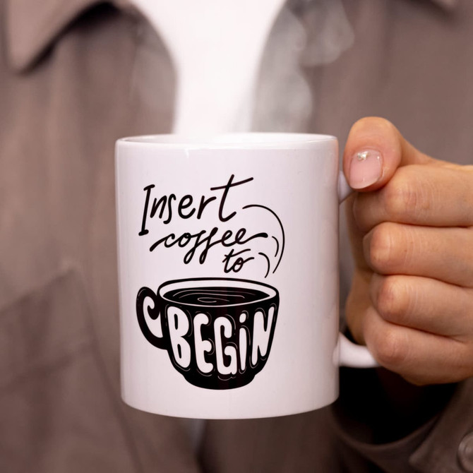 A white coffee mug with a design featuring a steaming cup and text, held by a person.