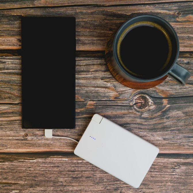 A 10,000mAh power bank in white, connected to a smartphone, is placed on a wooden surface alongside a black coffee mug.