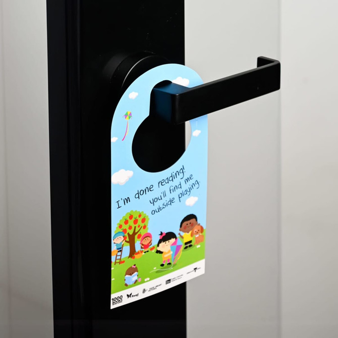 A double-sided door hanger featuring a colourful design with children playing outdoors, attached to a door handle.