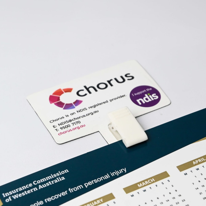 A white T-magnet with a clip, featuring a logo and contact information, holding a calendar.