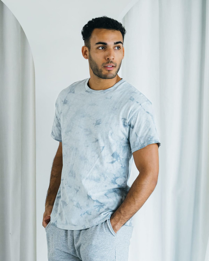 A man wearing a grey tie-dye t-shirt and light grey shorts, standing against a soft, neutral background.