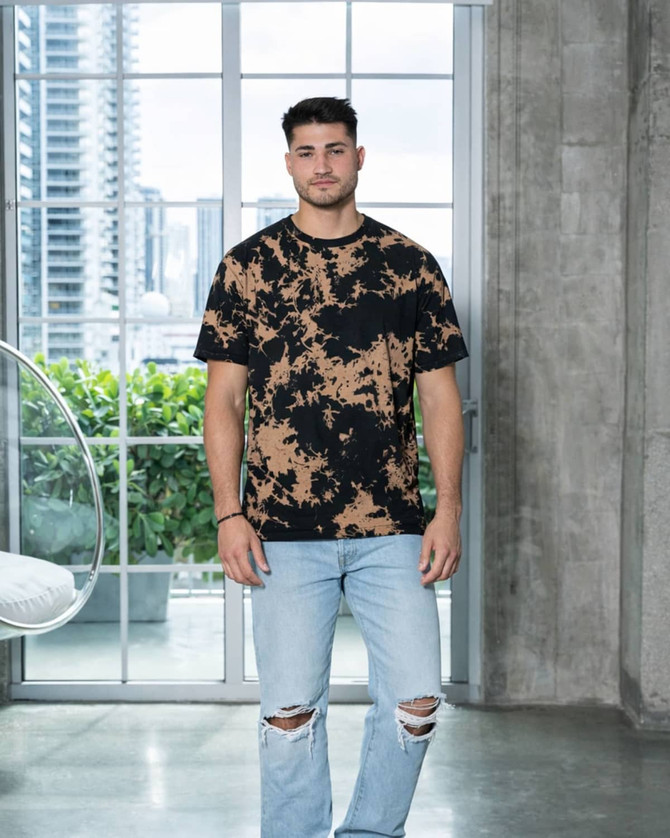 A man wearing a black and tan bleach-dyed T-shirt and distressed denim jeans, standing in a modern interior.