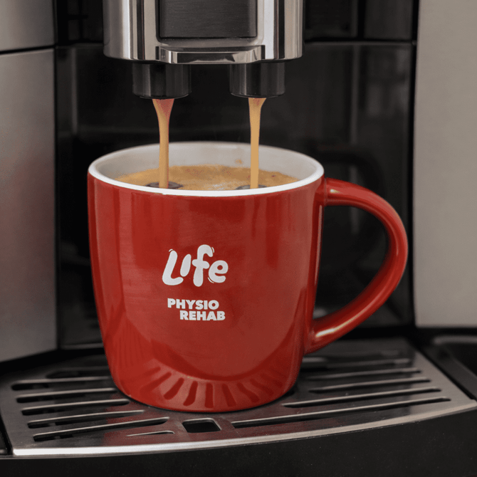 A red ceramic mug with a handle, featuring a logo, is filled with coffee under a machine.