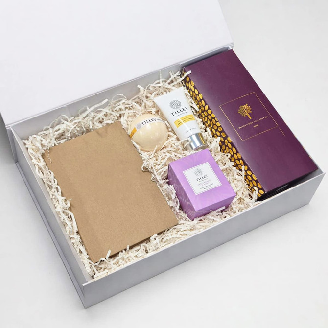 A gift box containing a brown notepad, a round item, a cream tube, a purple candle, and an item with a logo.