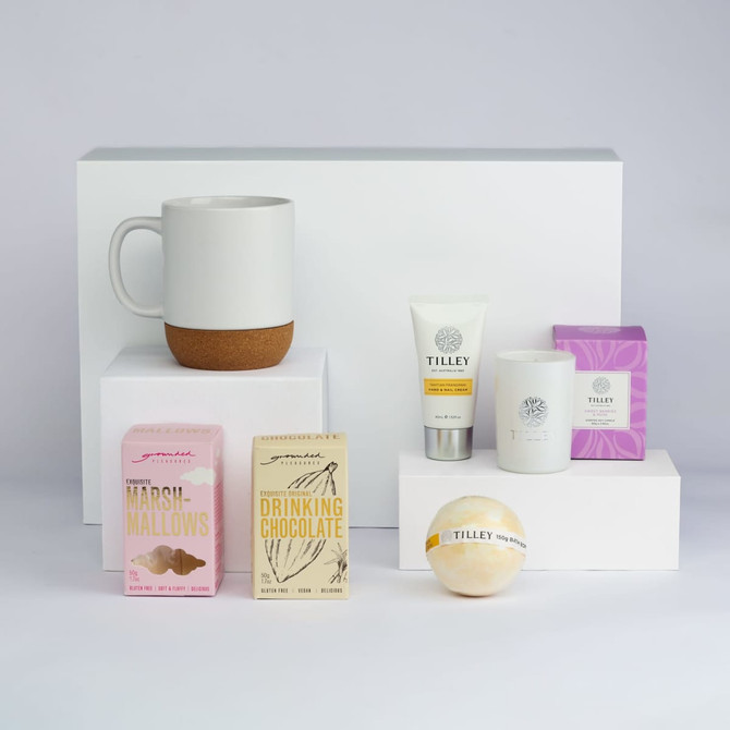 A white mug with a cork base, marshmallows, drinking chocolate, a candle, body cream, and a bath bomb in various packaging.