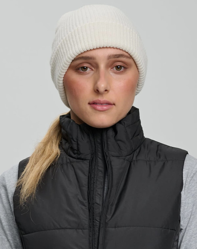 A person wearing a cream-coloured roll-up cable knit snug beanie with a black vest over a grey top.