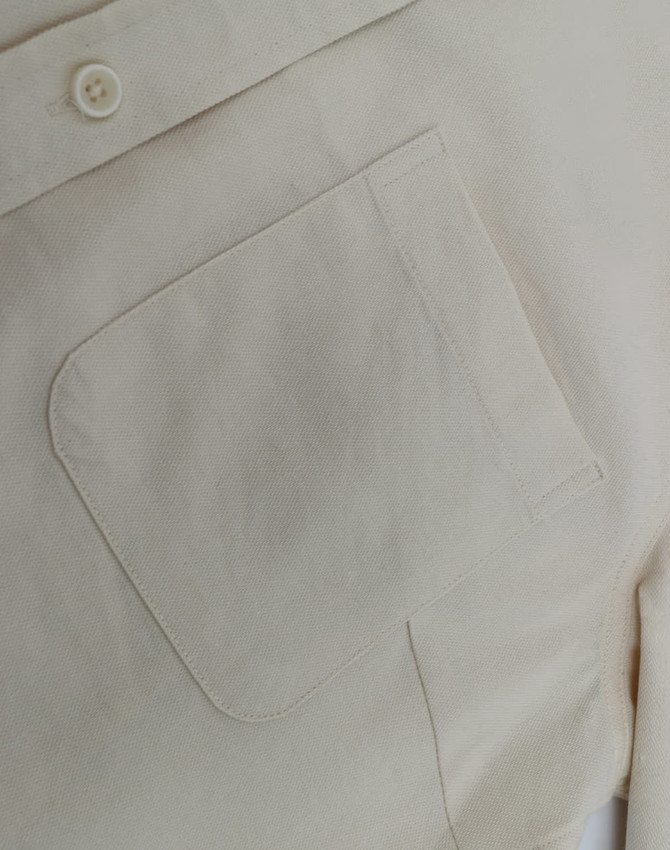 A close-up of a light beige cotton blend long-sleeve shirt, featuring a chest pocket and a button detail.