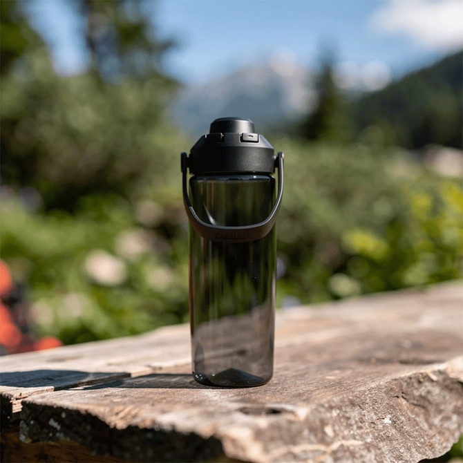 A drink bottle in a sleek, transparent design with a black lid and handle, set against a natural outdoor backdrop.