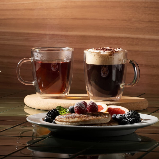 A set of two clear double wall mugs, one filled with coffee and the other with a dark beverage, alongside a plate of pancakes and berries.