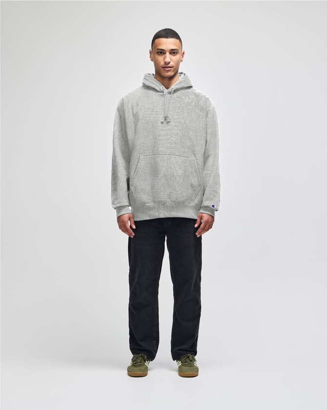 A man wearing a light grey hoodie with a hood, paired with dark pants, standing against a plain background.