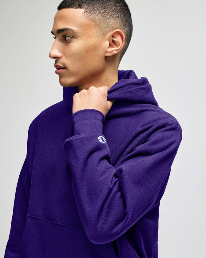 A purple hooded sweatshirt with a logo on the sleeve, worn by a model. The sweatshirt features a high neckline.