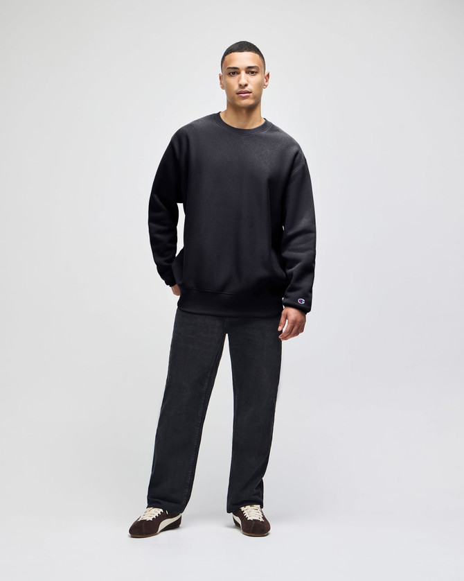 A black crew sweatshirt displayed on a model paired with dark pants and sneakers, featuring a logo.
