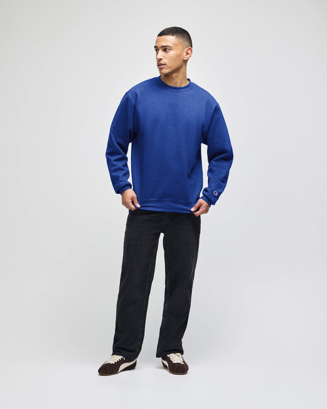 A blue crewneck sweatshirt with long sleeves, styled with dark trousers, featuring a logo on the sleeve.