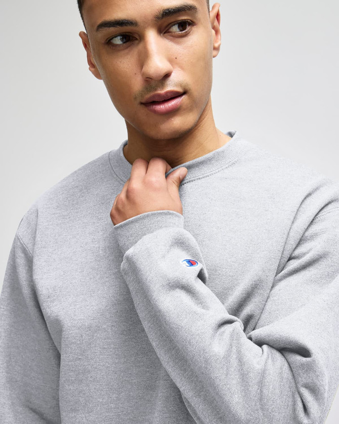 A grey Champion Powerblend® adult crewneck sweatshirt with a logo detail on the sleeve, displayed by a model.